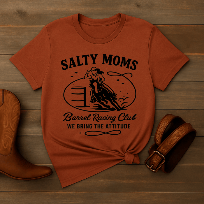 Orange t-shirt with 'Salty Moms Barrel Racing Club' design on a wooden surface with cowboy boots and a belt.