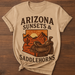 Beige t-shirt with 'Arizona Sunsets & Saddlehorns' graphic on a wooden surface