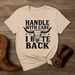 Beige t-shirt with graphic design and text on a wooden surface with boots and a belt.
