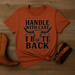 Orange t-shirt with graphic design and text on a wooden surface with cowboy boot and belt.