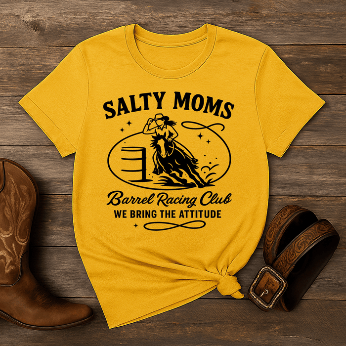 Yellow t-shirt with 'Salty Moms Barrel Racing Club' design on a wooden surface with cowboy boots and a belt.
