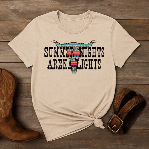 Beige t-shirt with 'Summer Nights Arena Lights' design on a wooden surface with cowboy boots and a belt.