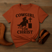 Orange t-shirt with 'Cowgirl for Christ' graphic on a wooden surface with cowboy boots and belt.