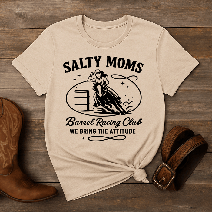Beige t-shirt with 'Salty Moms Barrel Racing Club' graphic on a wooden surface with cowboy boots and a belt.