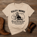 Beige t-shirt with 'Salty Moms Barrel Racing Club' graphic on a wooden surface with cowboy boots and a belt.